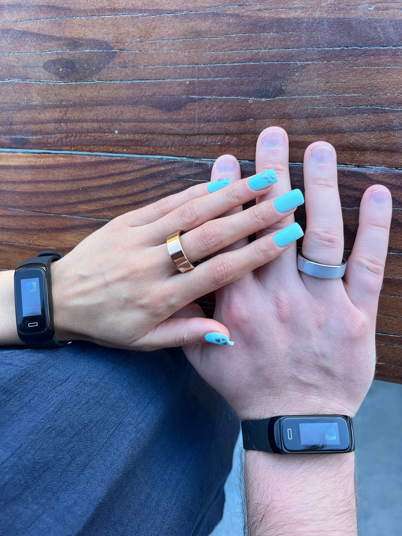 Two hands with nail polish, one wearing a ring and a fitness tracker, on a wooden surface.