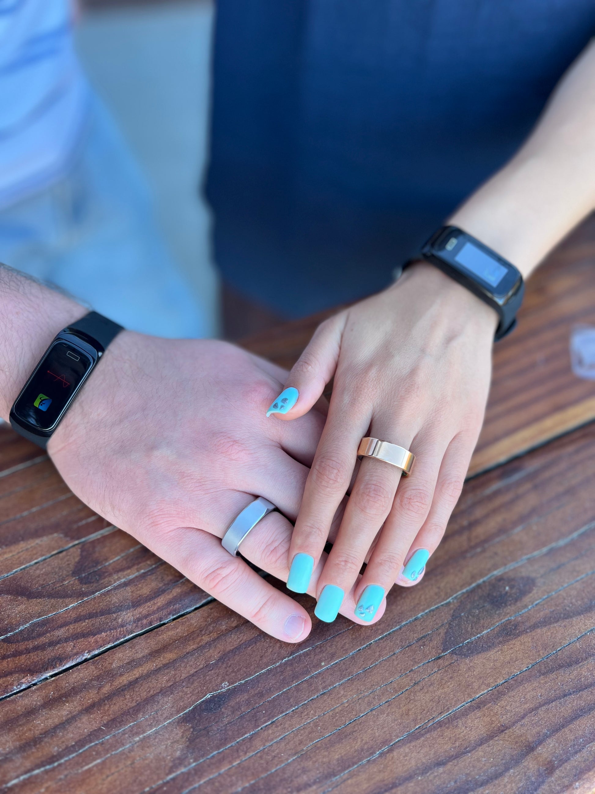 Two hands with rings on a wooden surface, one hand wearing a smartwatch.